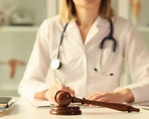 A doctor wearing a white coat sitting behind a witness stand.