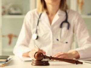 A doctor wearing a white coat sitting behind a witness stand.