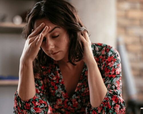 Woman sitting indoors with eyes closed and hand on forehead, showing signs of head pain