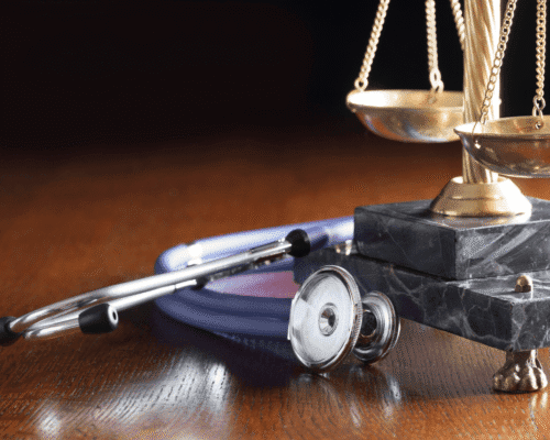 Close-up of a stethoscope next to brass legal scales on a polished wooden surface, symbolizing the intersection of healthcare and legal accountability