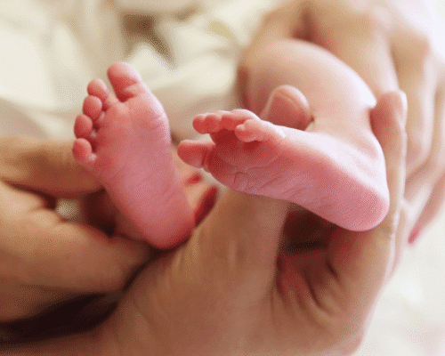 Close-up of an adult gently cradling a newborn’s small feet in their hands. The baby’s skin appears soft and pink, symbolizing care, tenderness, and the fragility of new life. The image conveys themes of parental love, loss, and medical care related to infant health.