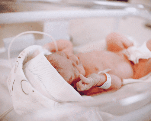 A newborn baby wearing a small white cap and hospital identification bracelet rests in an incubator. The baby’s delicate hand is slightly curled near its face, and soft lighting emphasizes the hospital environment. The image represents the topic of birth injury and medical care for newborns.