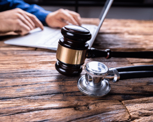 A judge’s gavel and a stethoscope rest on a wooden desk near a person typing on a laptop. The scene represents the legal process of filing a medical malpractice claim and the connection between healthcare and law.