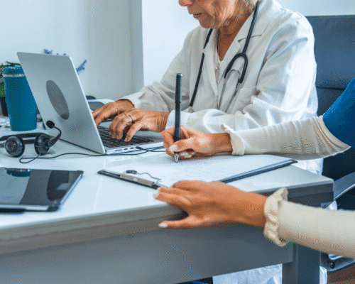 A doctor sits at a desk using a laptop while writing on a notepad, with a nurse assisting beside them. The image represents hospital record-keeping practices in Denver medical error cases, emphasizing the importance of accurate documentation for patient safety and malpractice investigations.